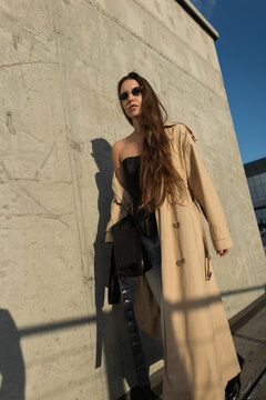 Low angle shot of a stylish woman in a beige trench coat and black corset, wearing sunglasses, posing near a concrete wall on a sunny urban platform.