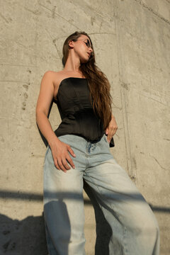 Confident young woman in a black satin corset top, wide-leg blue jeans, and sunglasses leaning on a sunlit concrete wall. Modern urban street style fashion.