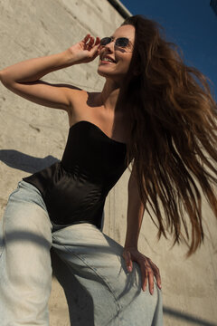 Sunny low-angle portrait of a confident young woman with braces and long, blowing hair, smiling up. She wears a black corset, jeans, and sunglasses against an urban concrete wall.