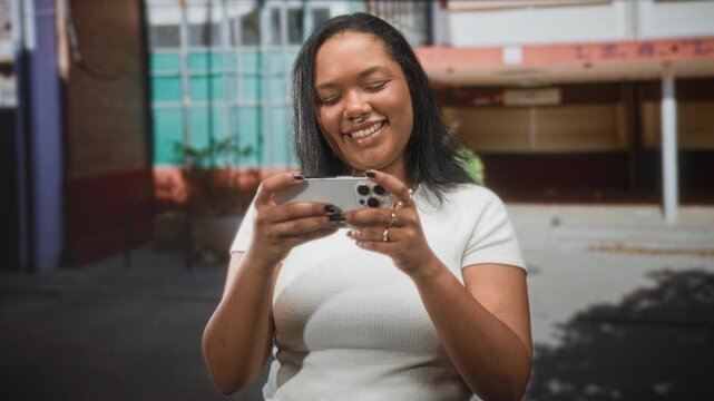 Young african american woman holding smartphone and smiling on a street in front of a building, thumb tapping screen; digital connection joy.