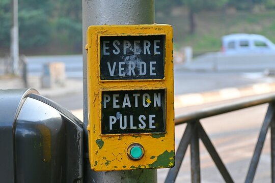 A close-up shot of a typical yellow pedestrian traffic light button in Madrid, Spain. The sign reads "Wait for green" and "Pedestrian, press",August 08, 2026