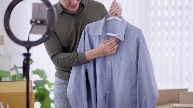 Young Asian online seller presenting blue shirt on hanger while filming product review in home studio