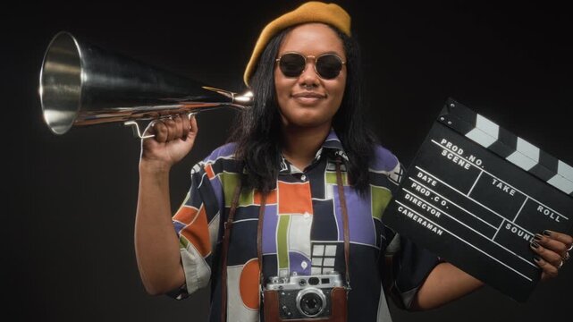 Woman holds clapperboard and metal megaphone with one hand, camera hangs from strap, wearing beret and sunglasses while smiling in studio; confidence creativity.
