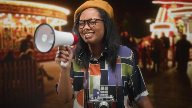 Young african american woman holding megaphone and wearing camera, speaking with closed eyes at a street carnival with carousel lights; creative celebration.