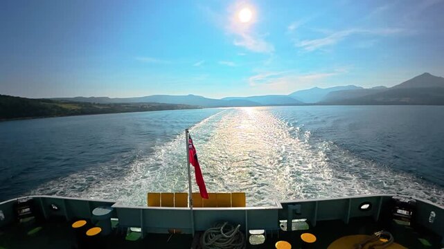Ferry boat creating a wake on the water, traveling towards distant mountains under a blue sky