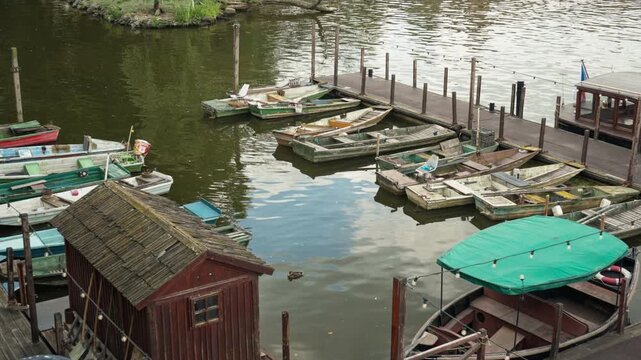 Wooden rowing boats moor at a pier in a river harbor. A small wooden shed stands on the quay for boat rental services. Traditional wooden vessels rest on calm water in a historic city berth.