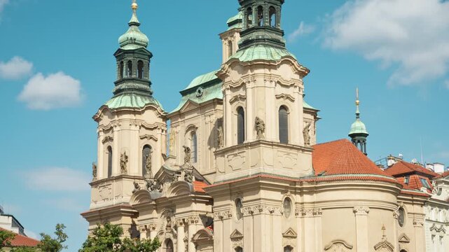 The facade of St. Nicholas Church stands in Prague. Ornate Baroque architecture features green domes and statues under a blue sky. Historic landmark attracts tourists in the old town square.