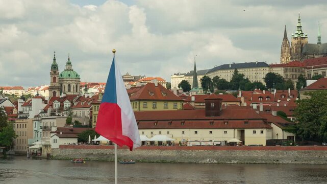 The Czech national flag flutters in the wind against the historic Prague skyline. Scenic city panorama features the Vltava River, traditional red roofs, and Prague Castle on a summer day.