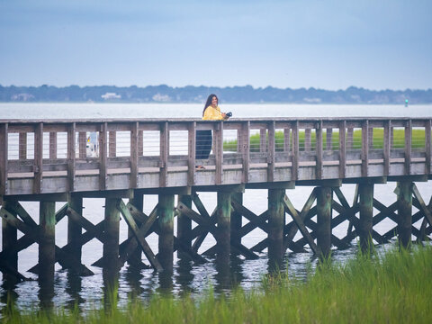 Female Photographer in Yellow Sweater Standing on Waterfront Dock Surrounded by Lowcountry Landscape at Sunrise in South Carolina
