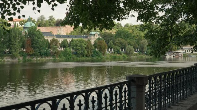 The Vltava River flows through Prague near a scenic promenade. A wrought iron fence lines the embankment under green trees. Historic architecture stands across the water on a bright summer day.