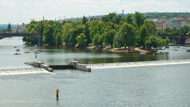 A low river weir spills water on the Vltava River near a green island in historic Prague, Czech Republic. Tourists ride pedal boats on calm water near a bridge on a sunny summer day.