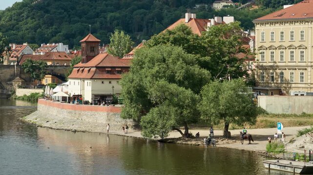 A scenic river embankment stretches along the Vltava River in historic Prague, Czech Republic. Green trees shade the peaceful waterfront near classic red roof architecture on a sunny day.