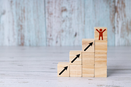 Concept of success, person reaching the top; wooden blocks or cubes arranged like a staircase, with an icon of a person raising their arms in a gesture of success at the top of the stairs.