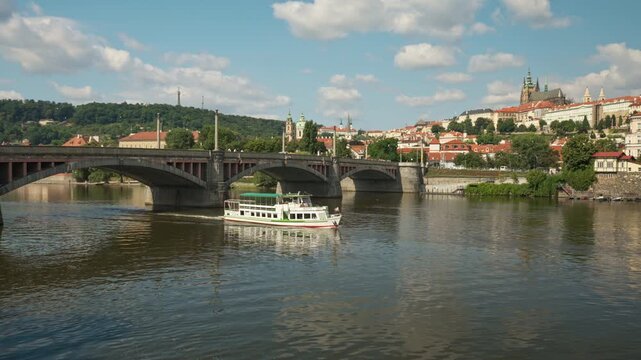A tourist boat cruises along the Vltava River in Prague. Historical architecture and Prague Castle stand in the background under a sunny sky. Excursion boat tours offer popular city travel.