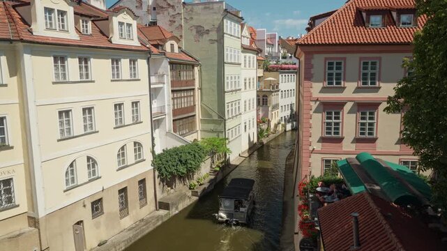 A small excursion boat sails down the narrow Certovka canal in Prague. Historic architecture frames the scenic Prague Venice waterway on a sunny day. Popular European tourist destination.