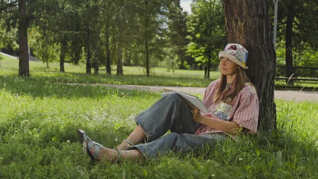 Caucasian young woman sitting on grass under tree in park, sketching in notebook and enjoying outdoor hobby. Footage is perfect for mental wellbeing campaigns and creative lifestyle ads