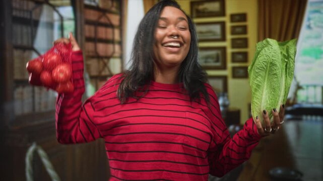 Young woman holding romaine lettuce and a bag of tomatoes, smiling and raising her hands in a building dining room; joy abundance.