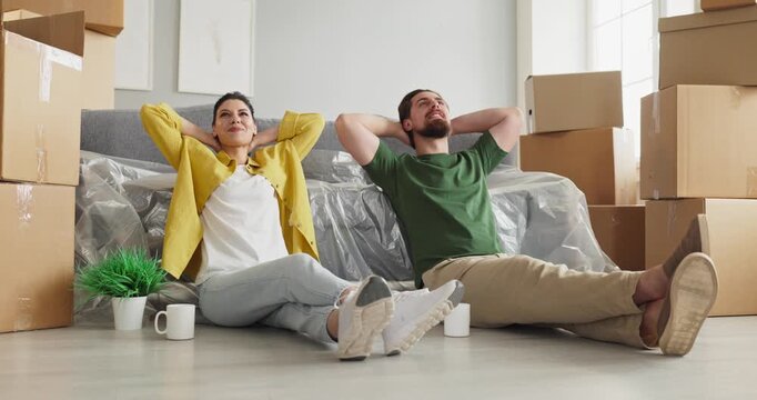 Couple moving unpacking relaxing boxes home new apartment. They lean back on the floor by a wrapped couch, smiling with mugs and a plant in bright daylight. Clear concept of moving and home