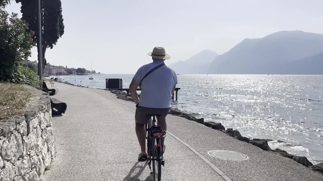 Man in blue shirt and straw hat cycling along Lake Garda in Malcesine, Italy