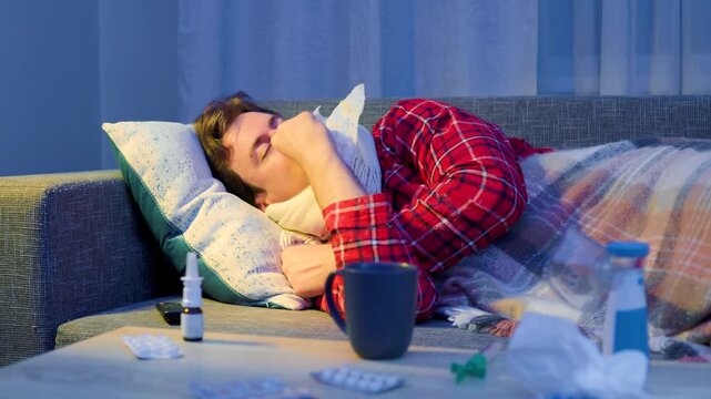 sick man with seasonal cold and flu symptoms rests on a couch under a blanket. He uses nasal spray for respiratory infection relief, practicing home treatment to recover. High quality 4k footage
