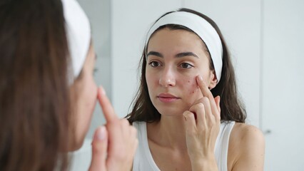 Fototapeta premium Young woman looking in mirror and touching acne on her cheek