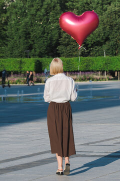 Beautiful blonde girl with short hair wearing a classic white blouse paired with a long brown skirt while holding a large, shiny red heart-shaped balloon