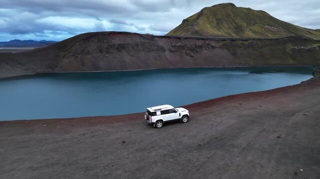 Aerial view of a white SUV parked on the volcanic rim of Blahylur crater lake with deep blue water and rugged terrain in Hella, Rangarthing ytra, Iceland.