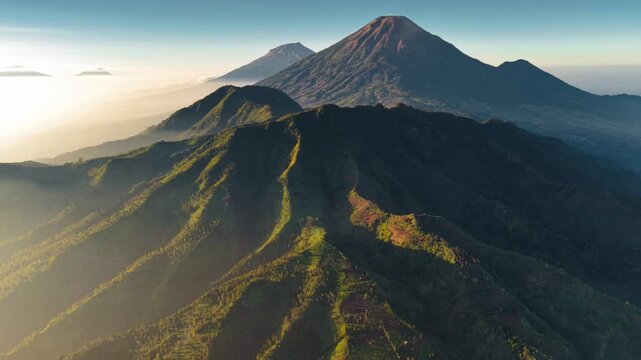 Aerial view of the volcanic landscape of Dieng Plateau with lush green ridges and distant peaks under a clear morning sky in Indonesia.