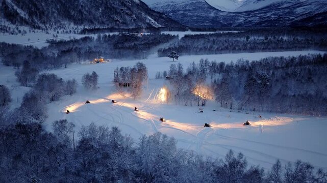 Aerial view of snowmobiles riding through snowy landscape at night with illuminated tracks in Tromso, Troms, Norway.