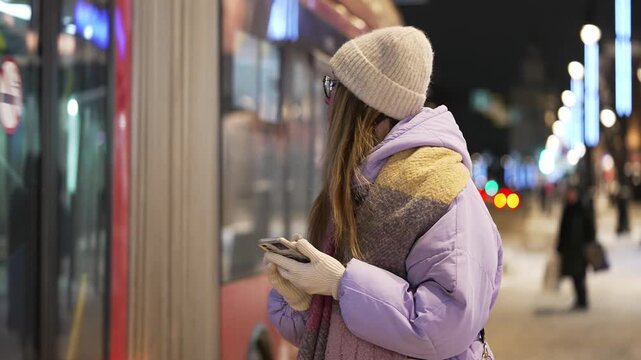 Young woman in a winter coat and hat using her mobile phone while waiting for public transport at a bus stop on a cold, snowy evening with city lights creating a beautiful bokeh in the background