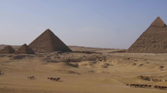 Panoramic view of the ancient Pyramids of Giza rising above the Egyptian desert, with camel caravans crossing the sandy landscape.