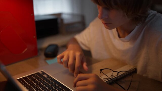 Focused teenager doing homework at home while working on a laptop and taking notes in a notebook. Schoolwork, studying, academic preparation and education concept.