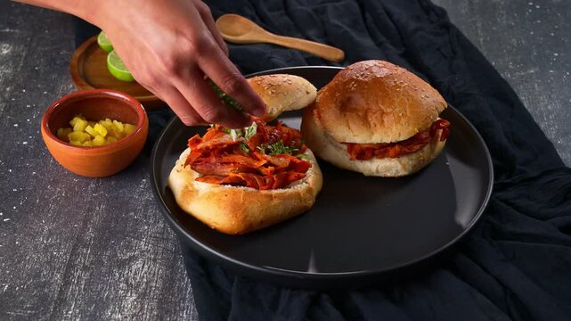 Person adding chopped cilantro and onion to traditional Mexican al pastor torta on black plate