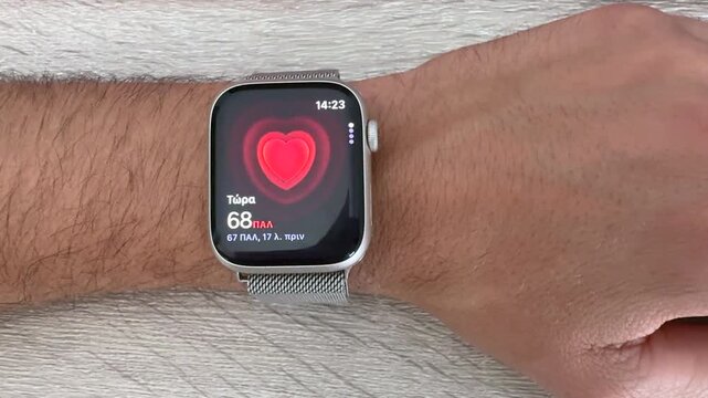 Close up shot of a person wearing a new smartwatch on their wrist displaying a red heart rate monitor interface while resting on a light wooden table highlighting modern health fitness technology.
