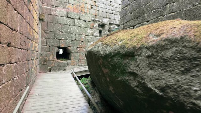 Point of view perspective moving backwards along a narrow wooden walkway between the ancient stone walls of a medieval fortress, showing the weathered texture of the rocks and moss