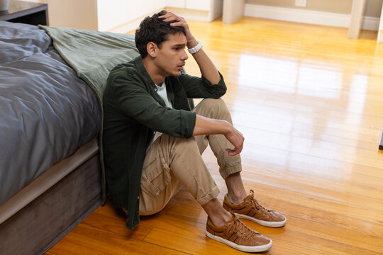African American man sitting on hardwood floor beside low platform bed, holding head, wearing watch