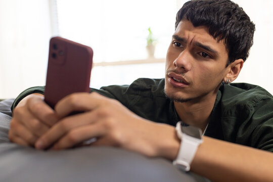 Adult man in dark green shirt leaning on gray couch studying red smartphone with white smartwatch