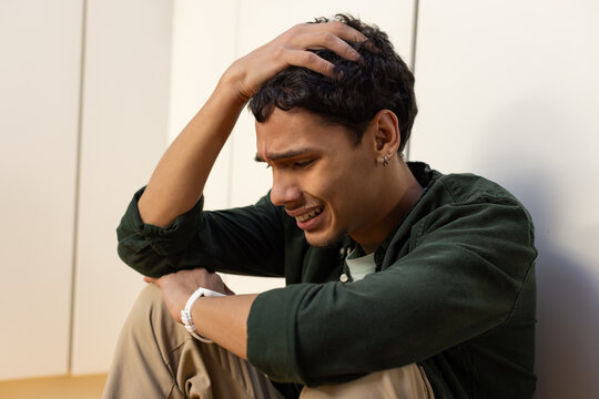 African American man sitting holding head against paneled wall, white watch and hoops, crying