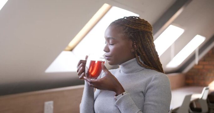 African woman holding glass mug, skylight drawing gaze, inhaling steam and sipping tea to relax