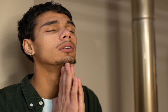 African man praying, hands at chin in hall, wearing shirt, hoop earring by metal post