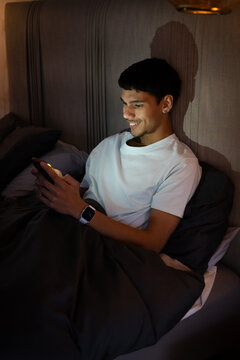 African American man in his early 20s holding smartphone and smartwatch while sitting up in bed