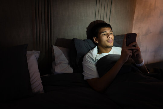 African American man reclining in bed at night, holding smartphone, wearing white tee, copy space