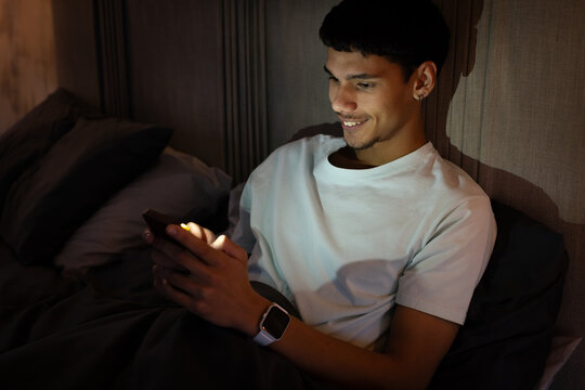 African American man sitting in bed at night holding phone with white tee smartwatch pillows duvet