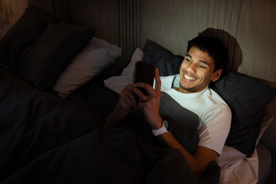 Adult African American male lying in bed at night holding smartphone with red case, copy space