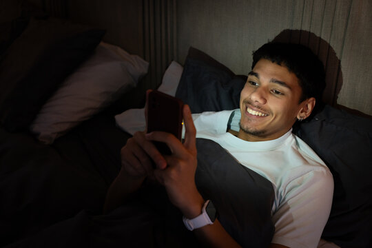 African American man reclining in bed at night holding lit smartphone, smiling and focusing