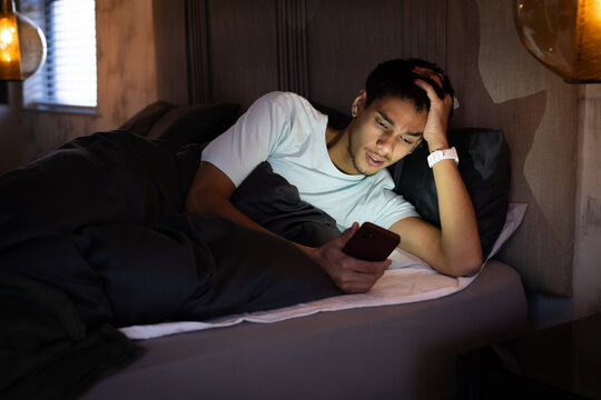 Man lying on bed at night holding smartphone, wearing white t-shirt and wristwatch