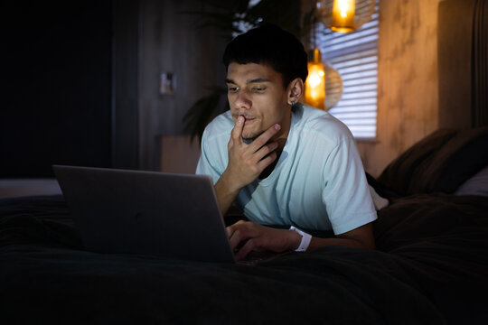 African American man lying on bed, propping on elbows, looking at laptop by pendant lamp