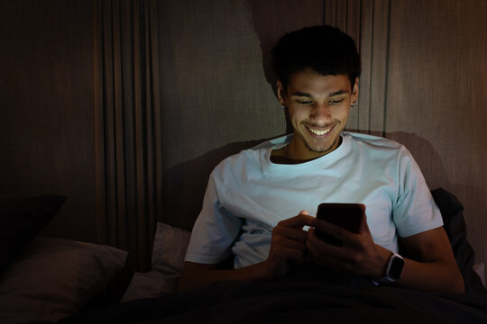 African American man in his twenties smiling, looking at phone in bed at night, wearing smartwatch