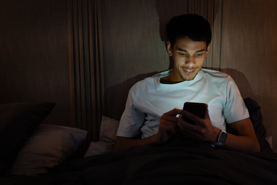 African American man reclining against headboard in bed, holding smartphone and wearing smartwatch