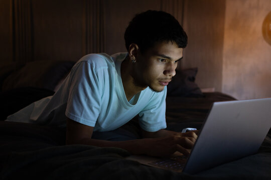 African American man in twenties lying on bed wearing light tee, focusing on laptop, holding phone